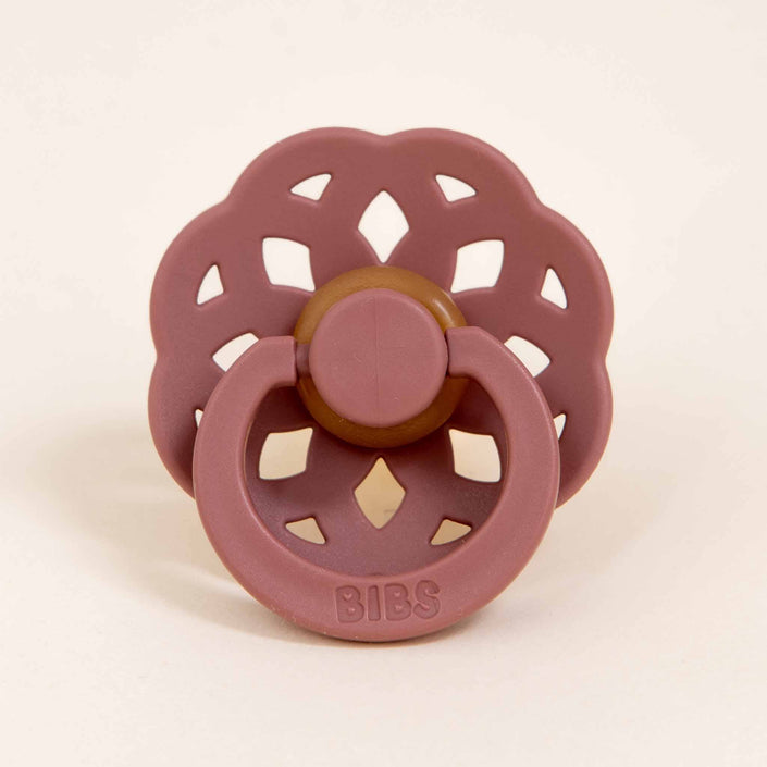 A pink Bibs Lace Pacifier from the Woodchuck 2 Pack, featuring a bohemian-inspired flower-shaped shield with a brown center, is placed on a light background. Made from natural rubber, the BIBS brand name is embossed on the handle, making it ideal for helping little ones self-soothe.