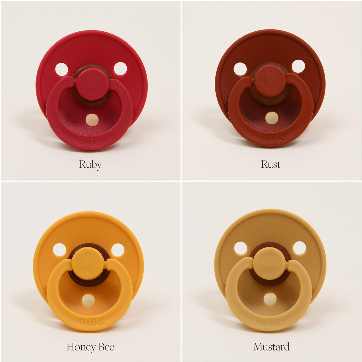 Displayed in a 2x2 grid are four Bibs Pacifiers, crafted from natural rubber. From top left to bottom right, the colors are labeled: Ruby, Rust, Honey Bee, and Mustard. Proudly made in Denmark, each pacifier includes a round shield and a ring handle to assist your baby in self-soothing with ease.