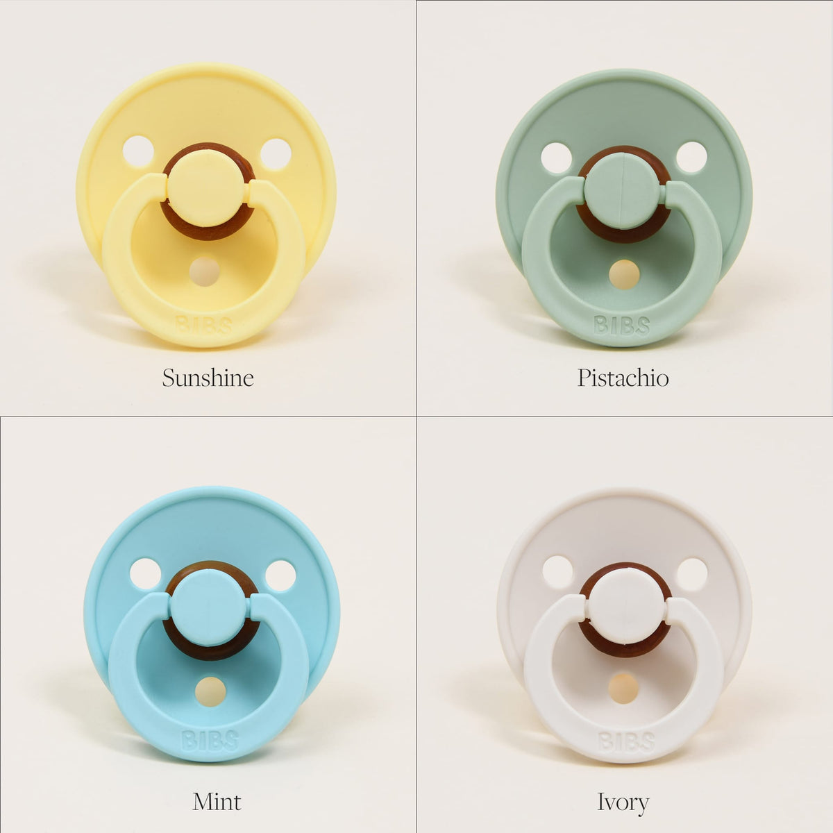A set of four Bibs Pacifiers crafted from natural rubber is arranged in a 2x2 grid on a light backdrop. The top left pacifier is yellow (Sunshine), the top right is green (Pistachio), the bottom left features a light blue hue (Mint), and the bottom right is white (Ivory). Designed in Denmark, these pacifiers offer babies a chic way to self-soothe.