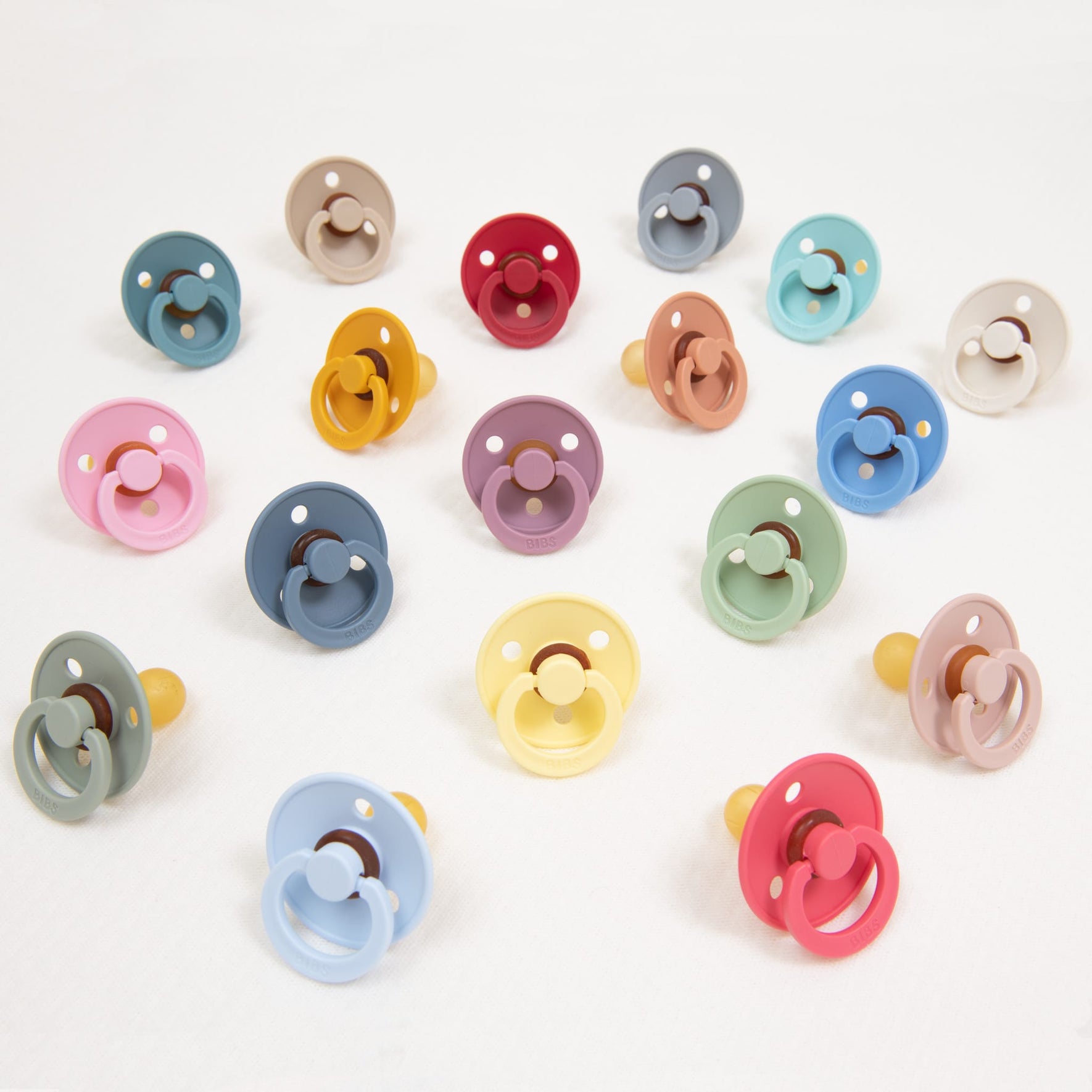 The image features a variety of colorful Bibs Pacifiers made from natural rubber, displayed on a white background. These pacifiers are available in vibrant shades such as pink, blue, green, yellow, and red. Manufactured in Denmark to assist babies in self-soothing, each pacifier includes a round shield with a circular handle and rubber nipple.