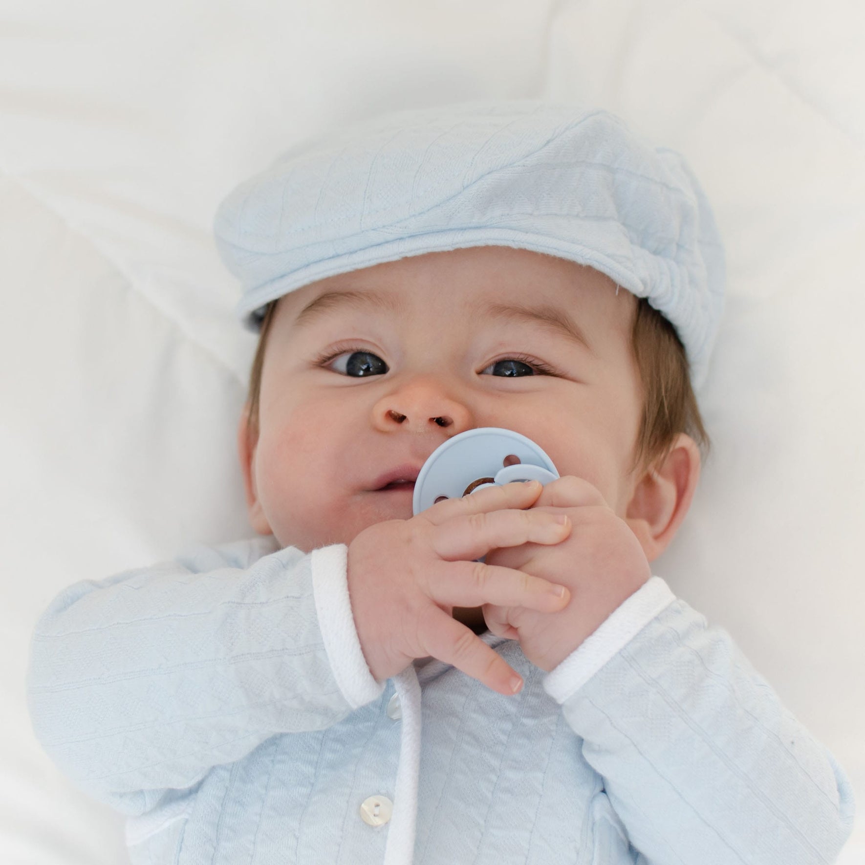 A baby in a light blue outfit and matching cap is lying on a white surface, calmly self-soothing with a Bibs Pacifier from the Baby Blue 2 Pack held close to its mouth.