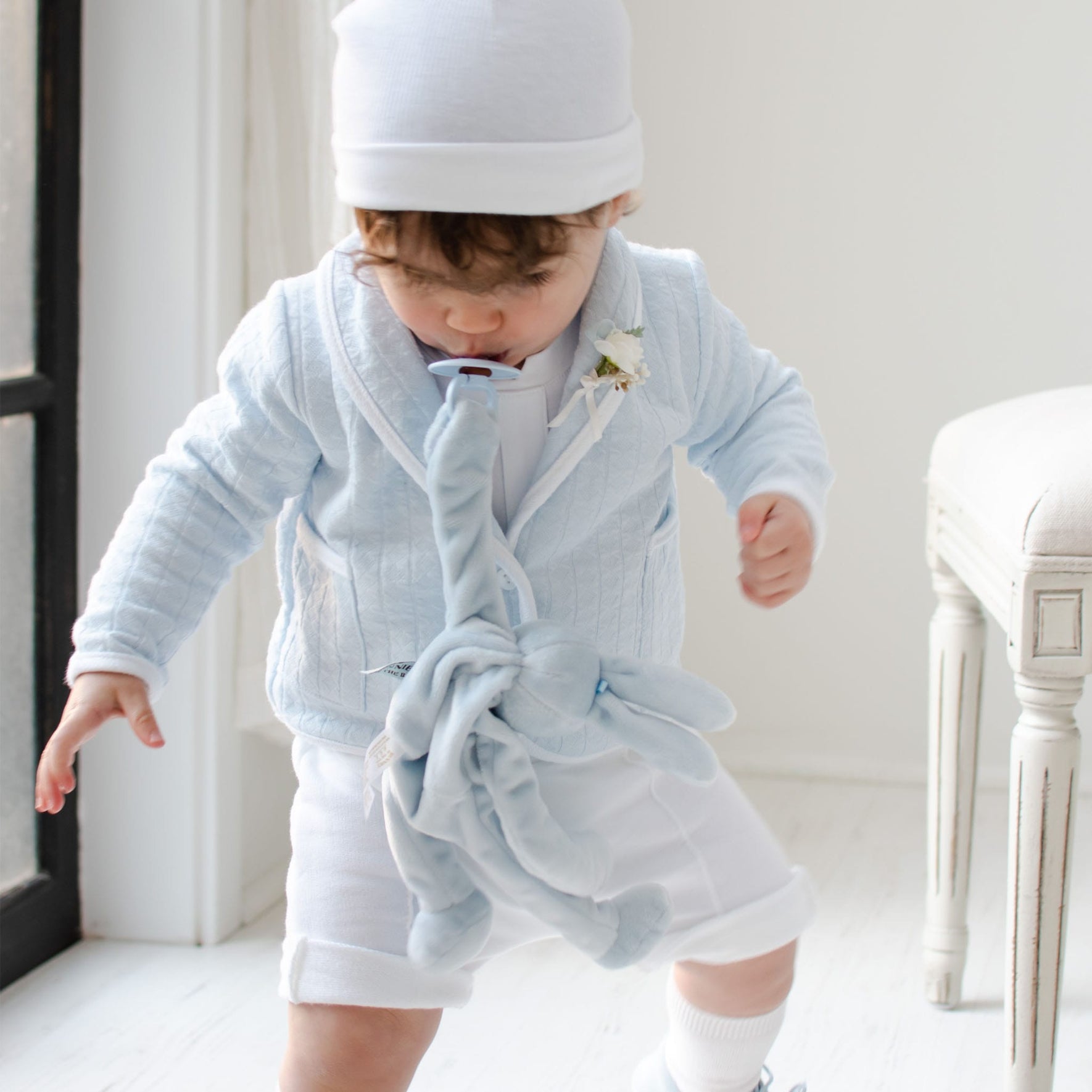 A toddler wearing a light blue outfit and matching hat stands indoors, holding a Bibs Pacifier in Baby Blue. The cozy room, adorned with white walls and light-colored furniture, exudes understated Scandinavian elegance reminiscent of Denmark's design ethos, creating a calming environment for the child to self-soothe.