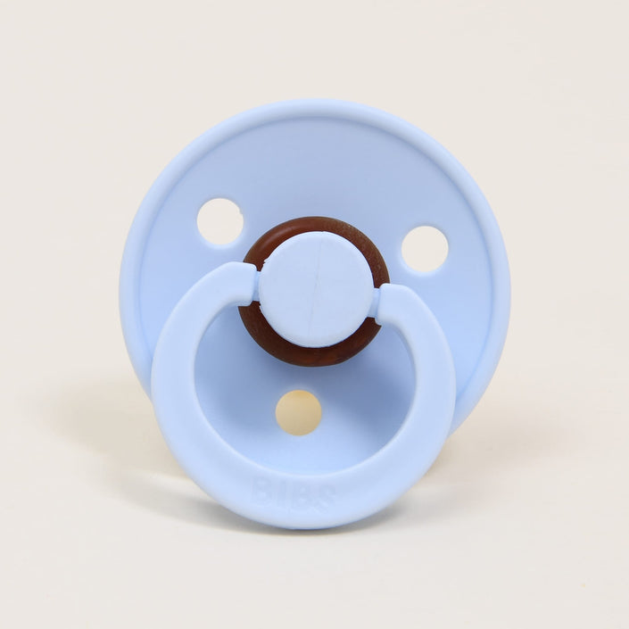 A baby blue Bibs pacifier from a 2-pack set, made of natural rubber with a round shield and handle, and crafted in Denmark. It is designed to assist babies in self-soothing, featuring three holes in the shield, and is displayed upright against a neutral background.