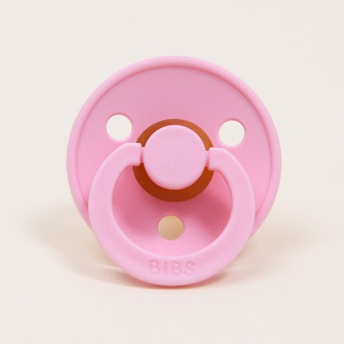 The Bibs Pacifier 2 Pack in Baby Pink showcases a classic design with a circular shield featuring two holes set against a plain white backdrop. The textured pattern on the shield enhances the embossed "BIBS" located near the base of the handle, crafted to aid babies in self-soothing.