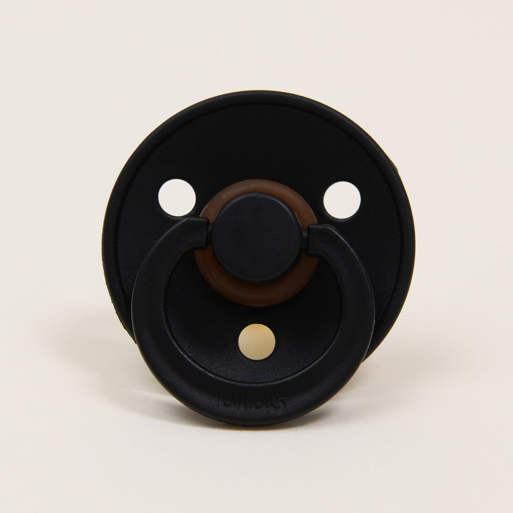 The Bibs Pacifier 2 Pack in Black showcases a sophisticated black pacifier with a brown center, made from natural rubber. Featuring three round holes in the shield, this elegant design originates from Denmark and provides soothing comfort for babies against a plain background.