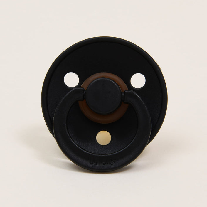The Bibs Pacifier 2 Pack in Black showcases a sophisticated black pacifier with a brown center, made from natural rubber. Featuring three round holes in the shield, this elegant design originates from Denmark and provides soothing comfort for babies against a plain background.