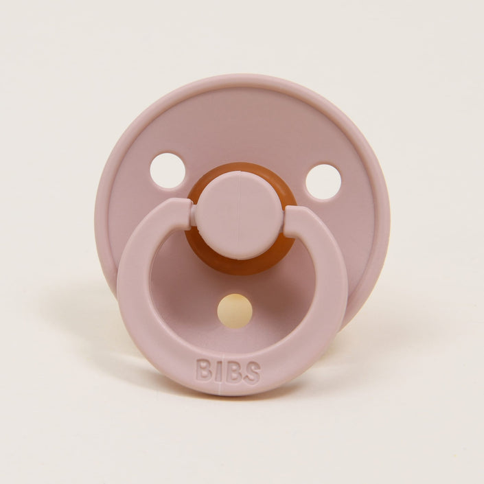 The Blush Bibs Pacifier from the 2 pack set is crafted from natural rubber and features a round shape with a circular handle. It includes three holes on its shield, with the word "BIBS" elegantly embossed on the handle, showcasing its quality craftsmanship from Denmark. Designed to help babies self-soothe, it rests upright against a light background.