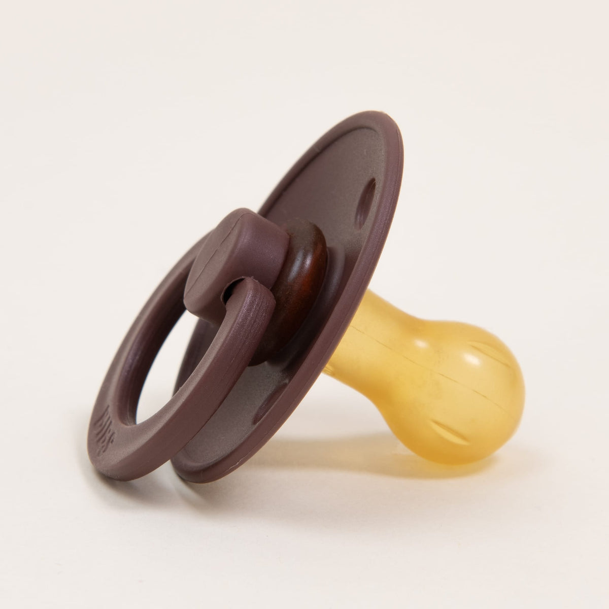 The Bibs Pacifier from the Chestnut 2 Pack, crafted from natural rubber, features a circular handle and nipple in shades of brown and yellow. Resting on a white surface, it boasts a straightforward yet functional design inspired by Danish craftsmanship, angled slightly upward to aid babies in self-soothing.
