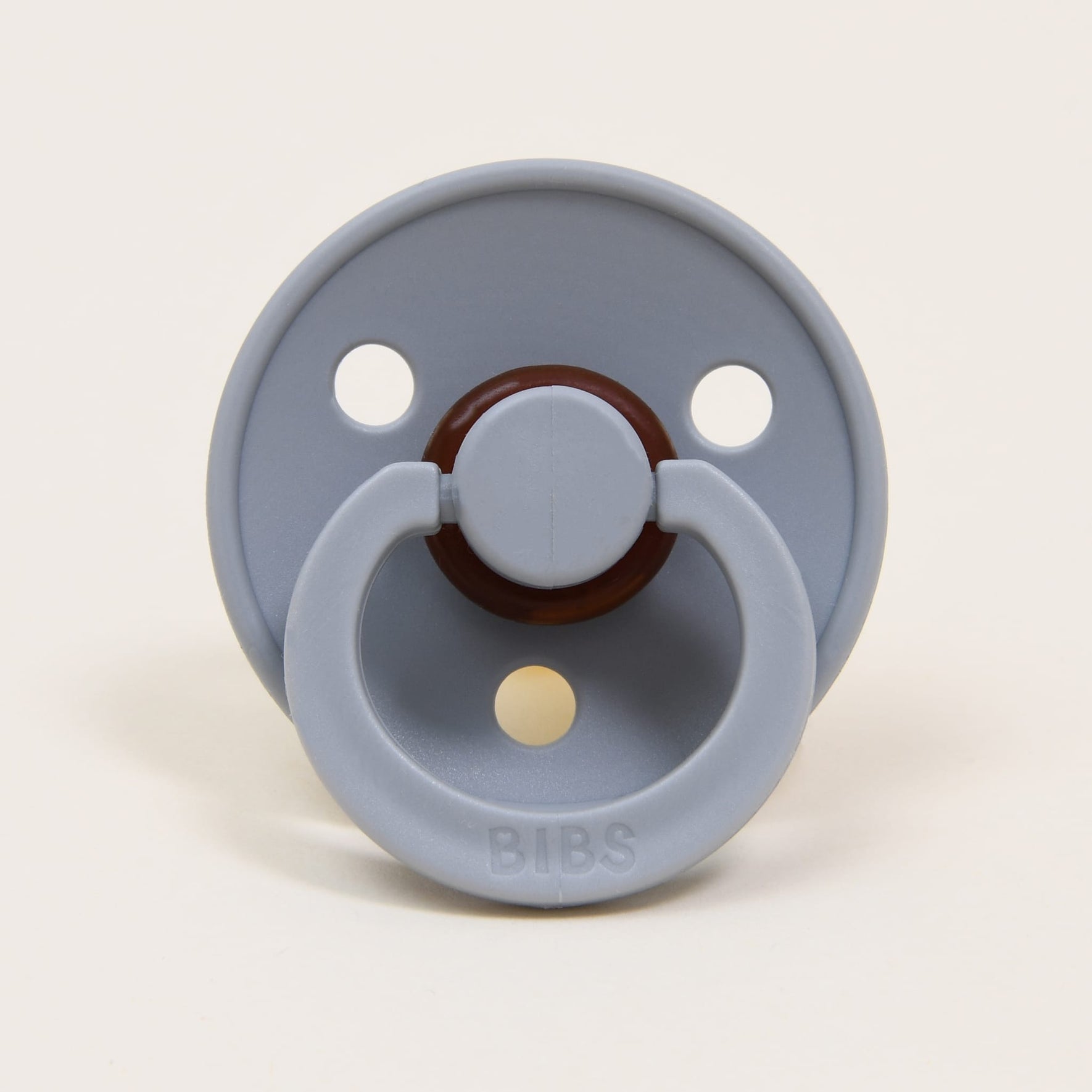 The Bibs Pacifier 2 Pack in Cloud is crafted from natural rubber and features a gray design with a round shield that includes three holes and a simple handle embossed with "BIBS." Made in Denmark, it is created to aid babies in self-soothing, with the off-white background accentuating its elegant appearance.