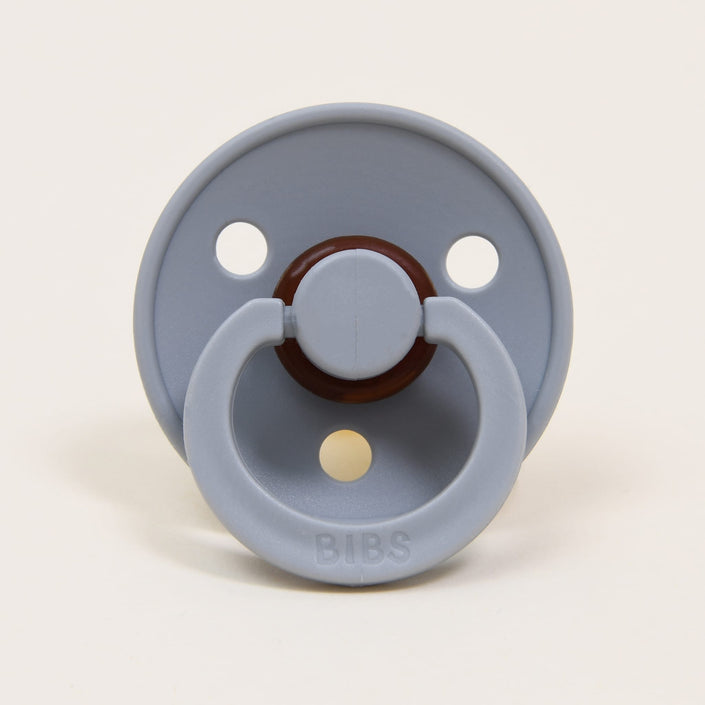 The Bibs Pacifier 2 Pack in Cloud is crafted from natural rubber and features a gray design with a round shield that includes three holes and a simple handle embossed with "BIBS." Made in Denmark, it is created to aid babies in self-soothing, with the off-white background accentuating its elegant appearance.