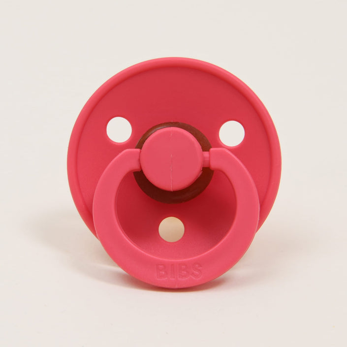 The Bibs Pacifier in Coral, expertly crafted in Denmark from natural rubber, features a circular shield with three ventilation holes and a looped handle, with "BIBS" embossed on the handle.