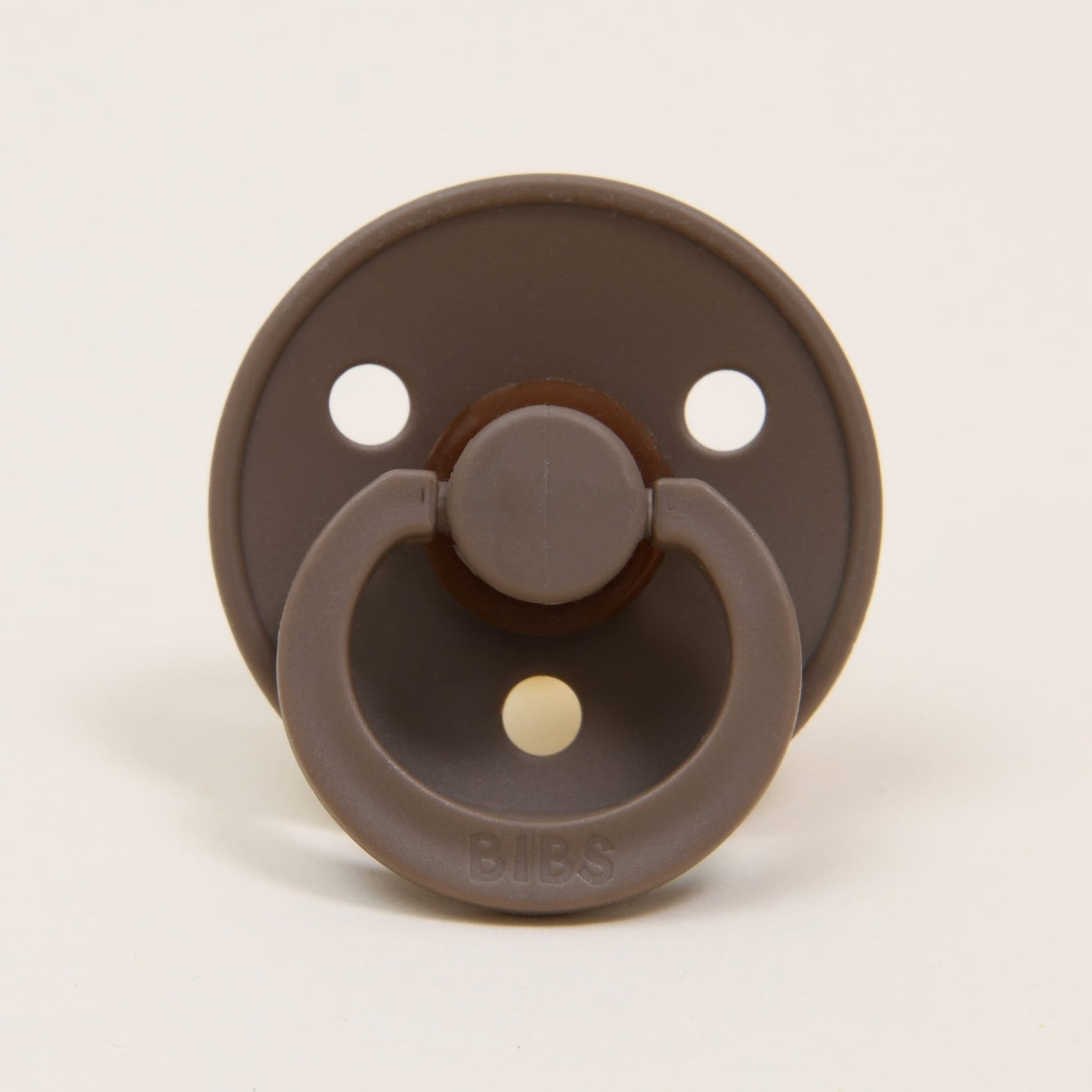 The Bibs Pacifier 2 Pack in Dark Oak showcases a classic design, with each pacifier featuring a natural rubber construction, a circular shield with three ventilation holes, and a ring handle. They are elegantly positioned upright on a neutral background to promote self-soothing.