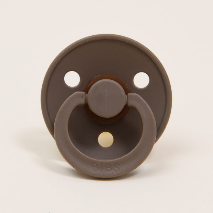 The Bibs Pacifier 2 Pack in Dark Oak showcases a classic design, with each pacifier featuring a natural rubber construction, a circular shield with three ventilation holes, and a ring handle. They are elegantly positioned upright on a neutral background to promote self-soothing.