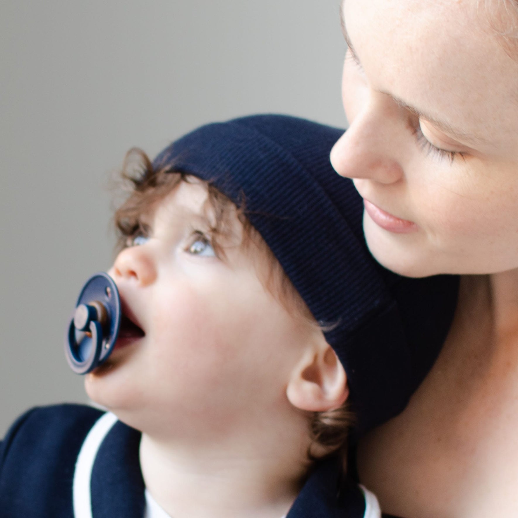 A woman with light skin gazes at a baby, who is self-soothing with a Bibs Pacifier from the Deep Space 2 Pack while wearing a navy blue hat. The baby's eyes wander upwards, dressed in navy blue clothing, as this tender moment unfolds against a softly blurred background.
