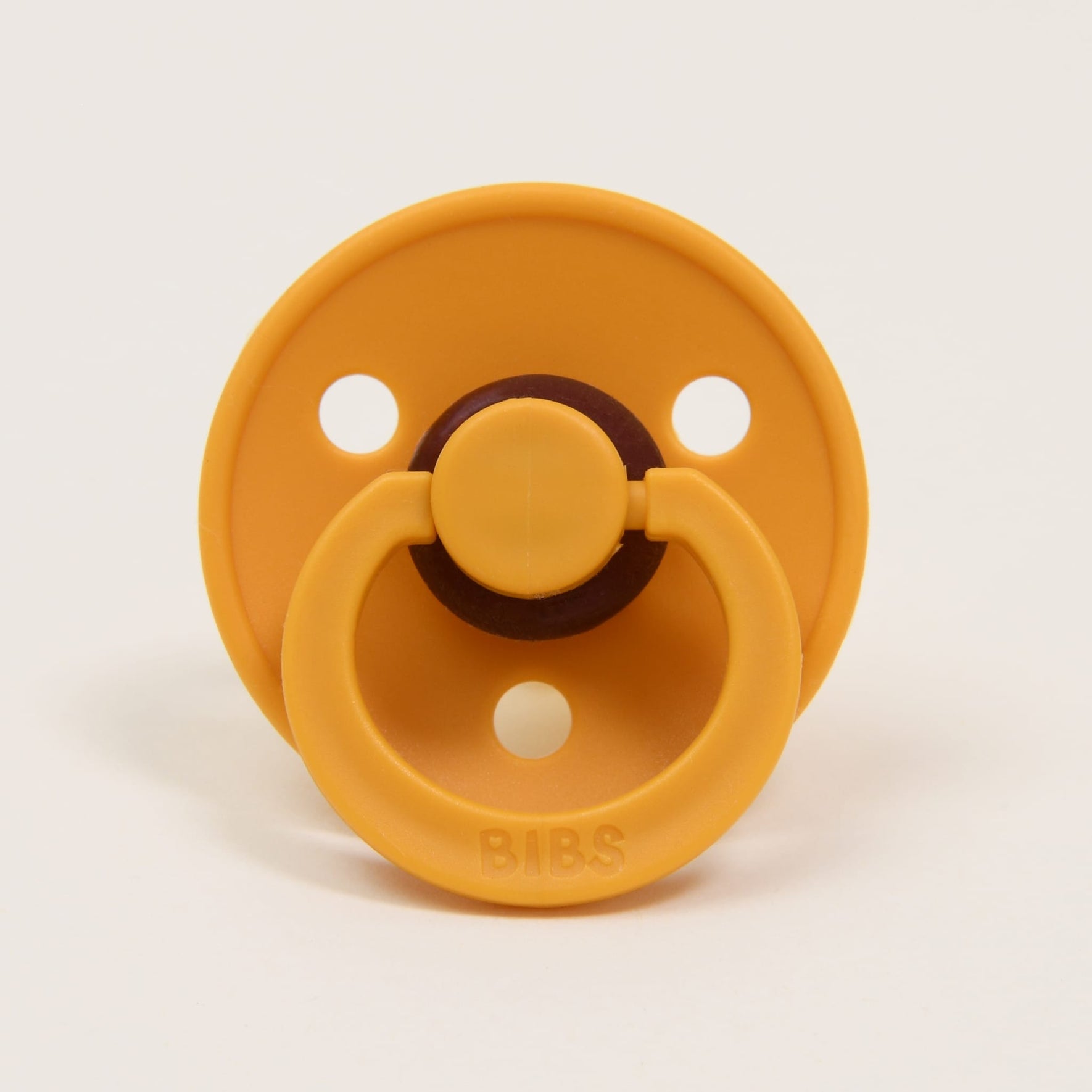 The Bibs Pacifier 2 Pack in Honey Bee is crafted from natural rubber and originates from Denmark. This cheerful yellow pacifier has a round shield with three ventilation holes. The front showcases a circular handle and features the "BIBS" branding embossed on its base, designed to help babies self-soothe against the simplicity of a plain white background.