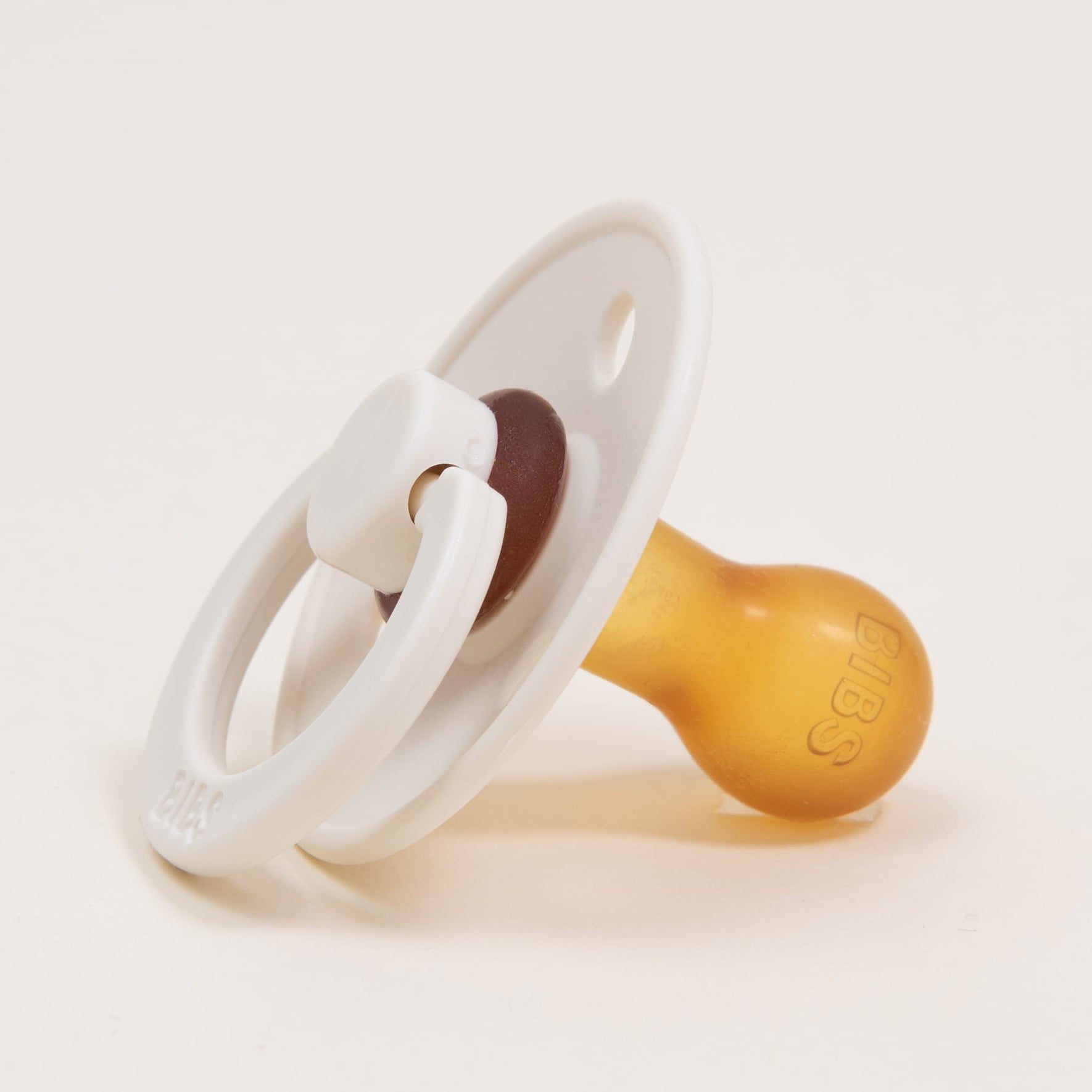 A timeless and elegant "Bibs Pacifier" from the 2 Pack in Ivory rests on a neutral surface, showcasing a sophisticated white and amber color scheme. It features a ring handle and round shield, with a natural rubber nipple ideal for self-soothing. The brand name "BIBS" is subtly embossed on its surface.