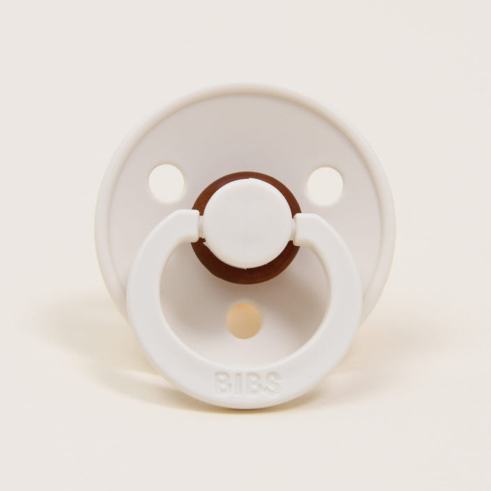 Among the showcased items, one is a Bibs Pacifier, known for its timeless elegance and crafted from natural rubber. It is displayed against a simple white backdrop, highlighting its ivory circular shield with two small openings at the top. The ring handle, embossed with "BIBS," is specifically designed to assist babies in self-soothing.