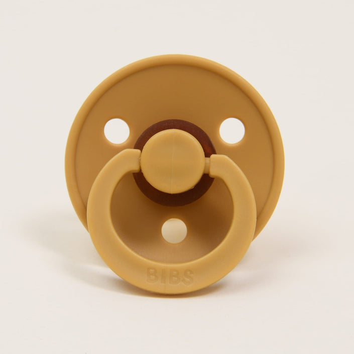 The Bibs Pacifier 2 Pack in Mustard features a classic design with a tan hue, round shield, and three holes. It includes a circular handle embossed with "BIBS," making it ideal for helping your little one self-soothe. The pacifiers are displayed upright against a plain white background.
