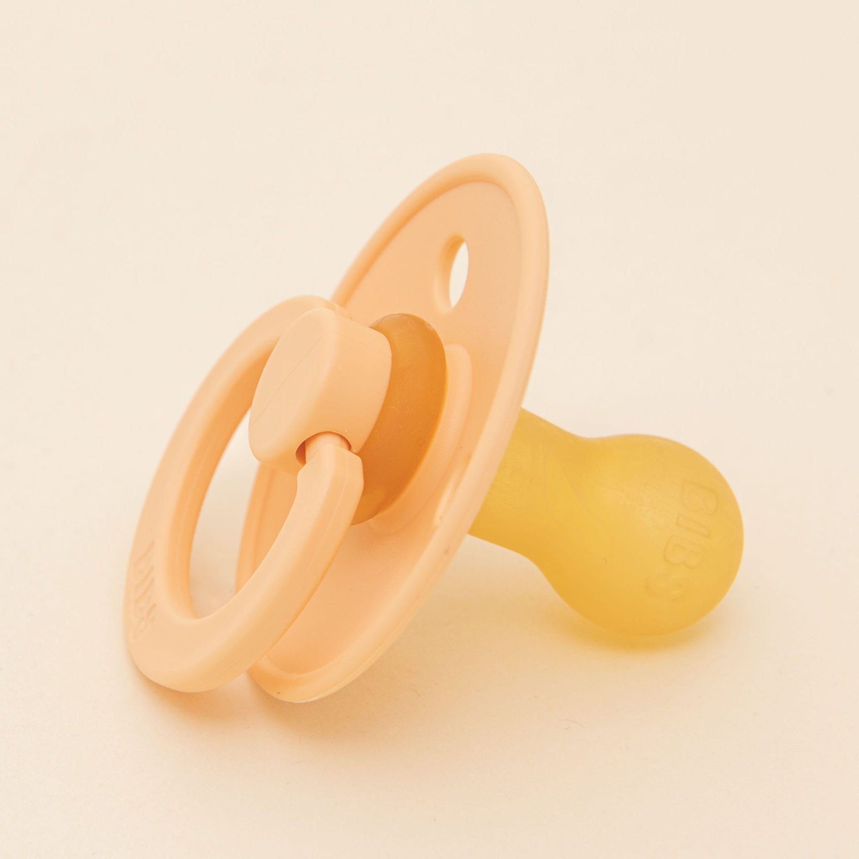 A soft, peach sunset-colored Bibs pacifier made from natural rubber, complete with a rounded nipple, delicately rests on a neutral background. This pacifier is designed to aid infants in self-soothing and showcases a circular handle attached to its shield, embodying the simplicity and craftsmanship synonymous with Denmark.