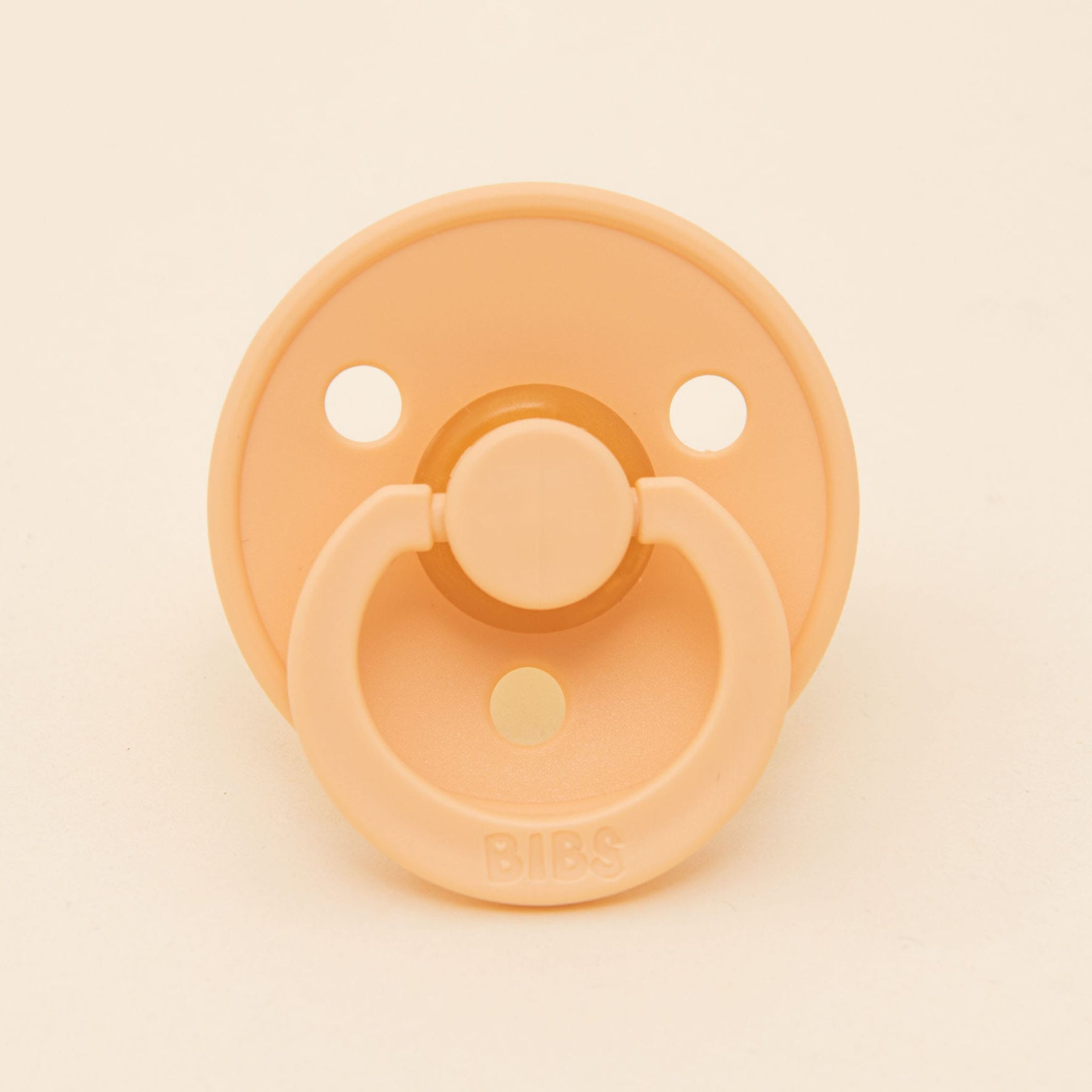 A Bibs Pacifier from the Peach Sunset 2 Pack, crafted from natural rubber, features a round shield with three ventilation holes and a ring handle embossed with the "BIBS" brand. This Danish-designed pacifier is ideal for helping little ones self-soothe, set against a light cream background.