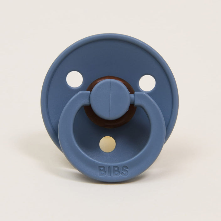 A Bibs Pacifier in Petrol, part of a 2 pack, is crafted from natural rubber and features two circular ventilation holes on the shield. The brand name "BIBS" is embossed on the handle. Designed to help babies self-soothe, it includes a round central part and a ring-shaped handle, all made in Denmark against a neutral background.