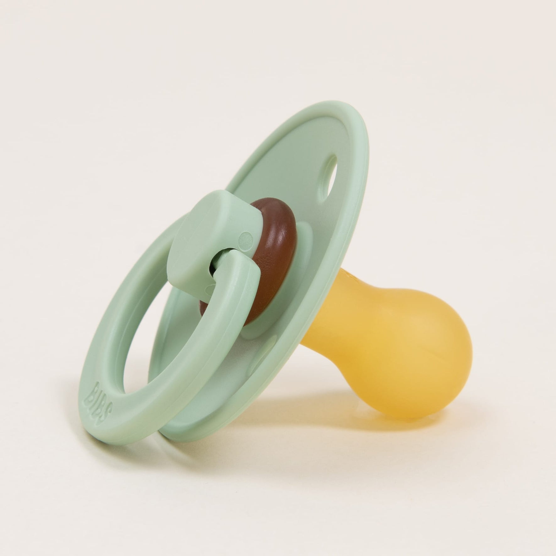 A Bibs Pacifier from the Pistachio 2 Pack, crafted from natural rubber, rests on a plain white surface. Its yellow nipple invites babies to self-soothe. Designed with a circular shield and handle, this Danish-inspired pacifier combines functionality and Scandinavian simplicity.