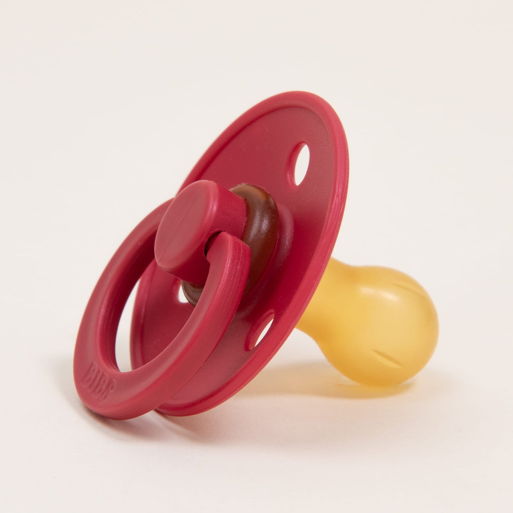 The Bibs Pacifier from the Ruby 2 Pack features a classic design in red and yellow, made from natural rubber with a round handle and two small holes on its shield. It's perfect for helping little ones self-soothe, all set against a plain white background.
