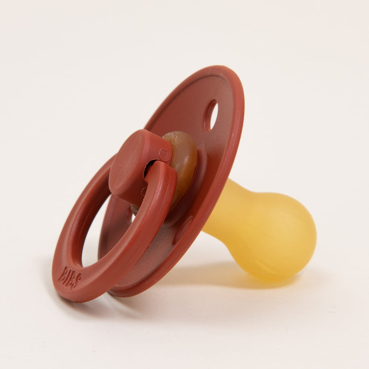 On a neutral background lies a timeless and elegant Rust Bibs Pacifier, crafted from natural rubber. It features a round shield and loop handle with a yellow nipple designed for soothing, embodying both simplicity and functionality.