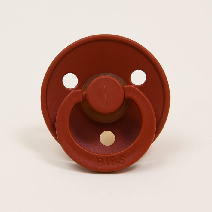 The Bibs Pacifier 2 Pack in Rust offers a timeless and elegant design, crafted from natural rubber. The red pacifier features three holes in its circular shield and a convenient ring handle. Set against a plain off-white backdrop, the handle is embossed with "BIBS," ready to soothe your little one.