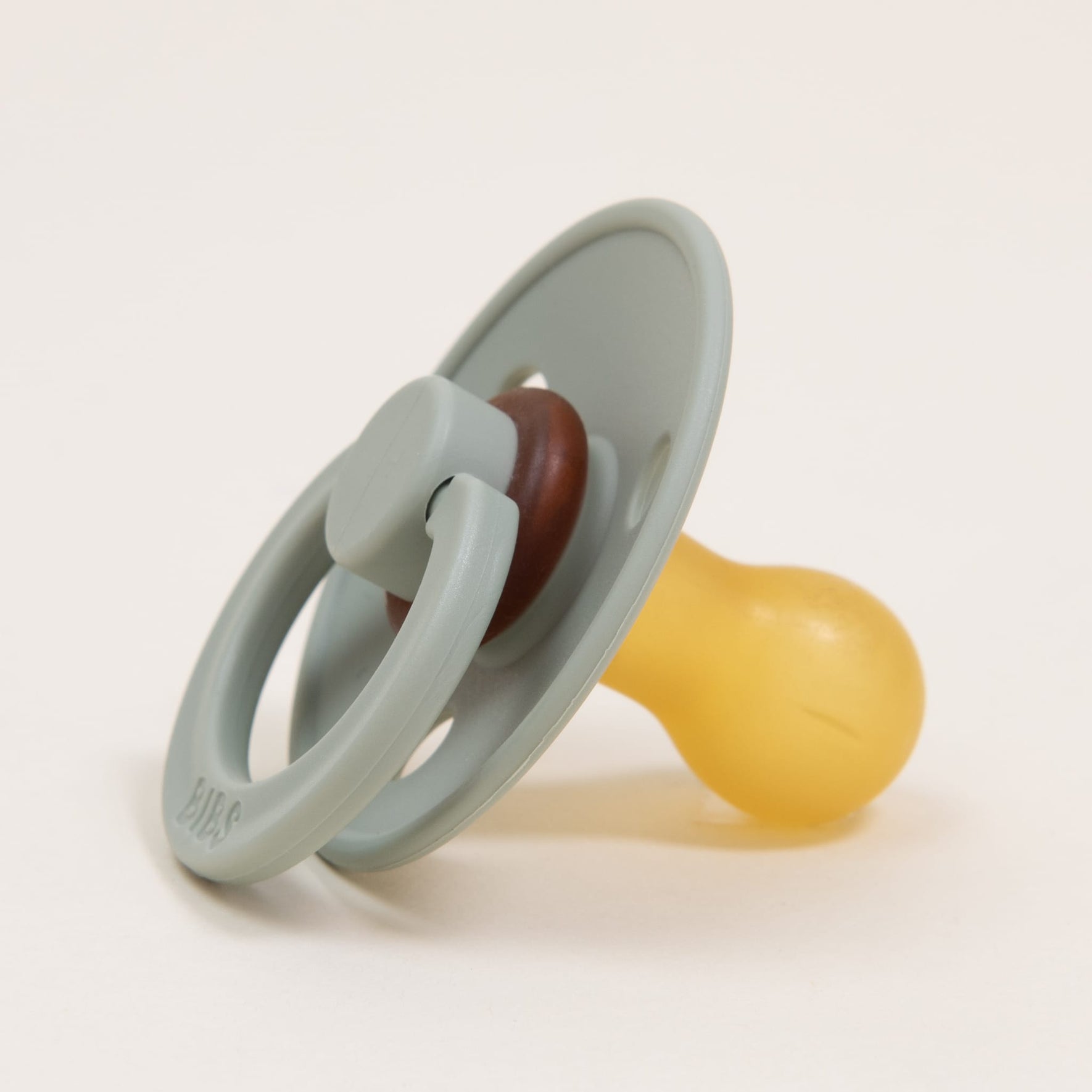 A single Bibs pacifier from the Sage 2 Pack, featuring a classic design with a sage green shield and a yellow natural rubber nipple, rests on a plain off-white background. Its timeless appearance and smooth finish offer an ideal solution for helping little ones self-soothe.