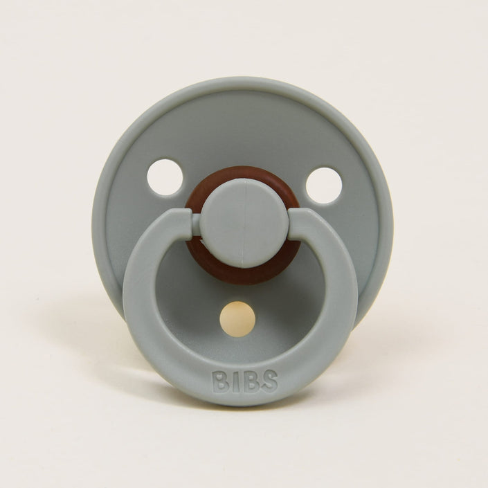 The Bibs Pacifier in Sage is elegantly designed with a gray tone and classic features, including two circular holes in the shield and a round, brown center crafted from natural rubber. The handle attached to the shield has "BIBS" embossed on it. Displayed upright against a white background, it is ideal for helping babies self-soothe.