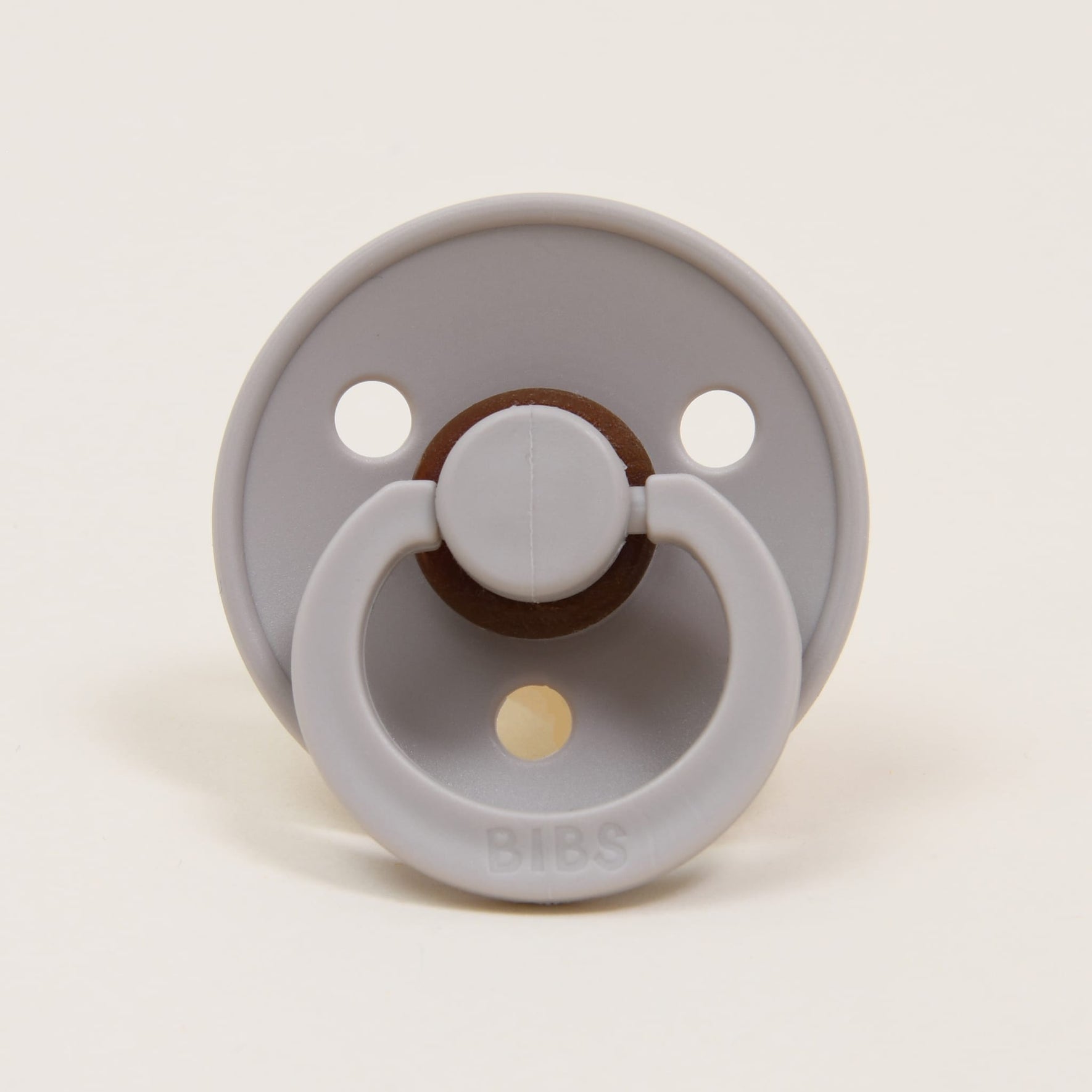 A sand-colored natural rubber pacifier from the Bibs Pacifier 2 Pack has a round shield with two small holes and a central looped handle. The brand name "BIBS" is embossed at the bottom. Made in Denmark, it provides a soothing touch for babies. The backdrop is a plain, light color.