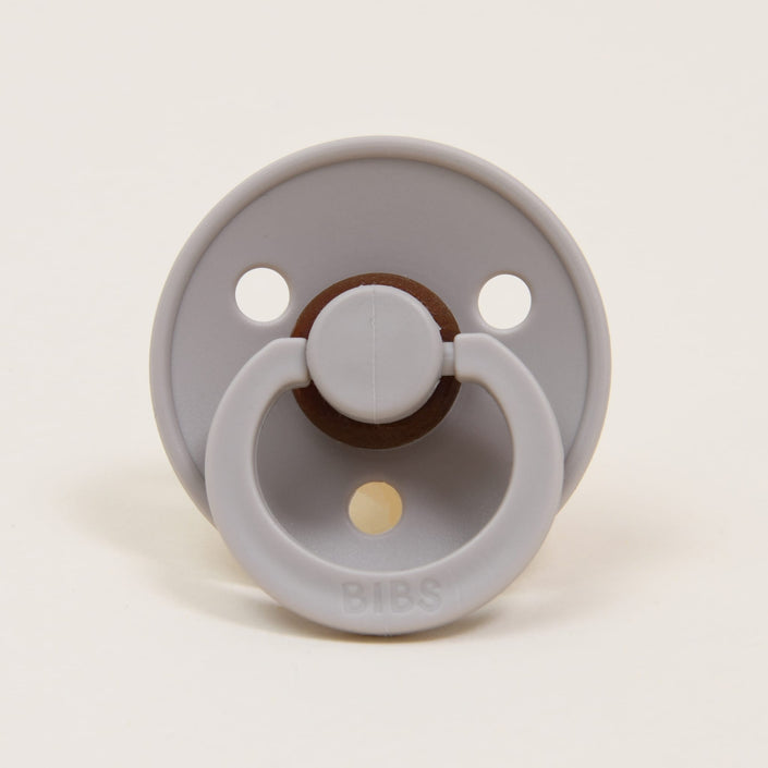 A sand-colored natural rubber pacifier from the Bibs Pacifier 2 Pack has a round shield with two small holes and a central looped handle. The brand name "BIBS" is embossed at the bottom. Made in Denmark, it provides a soothing touch for babies. The backdrop is a plain, light color.
