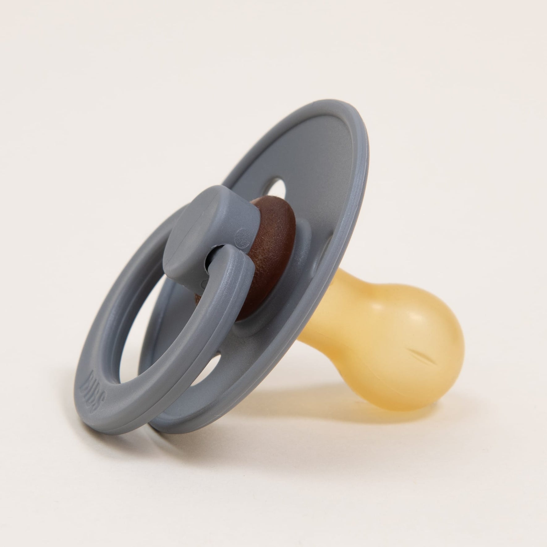A Smoke Bibs Pacifier, featuring a gray design with a circular handle and a beige silicone nipple made from natural rubber for babies to self-soothe, is placed on a neutral background.
