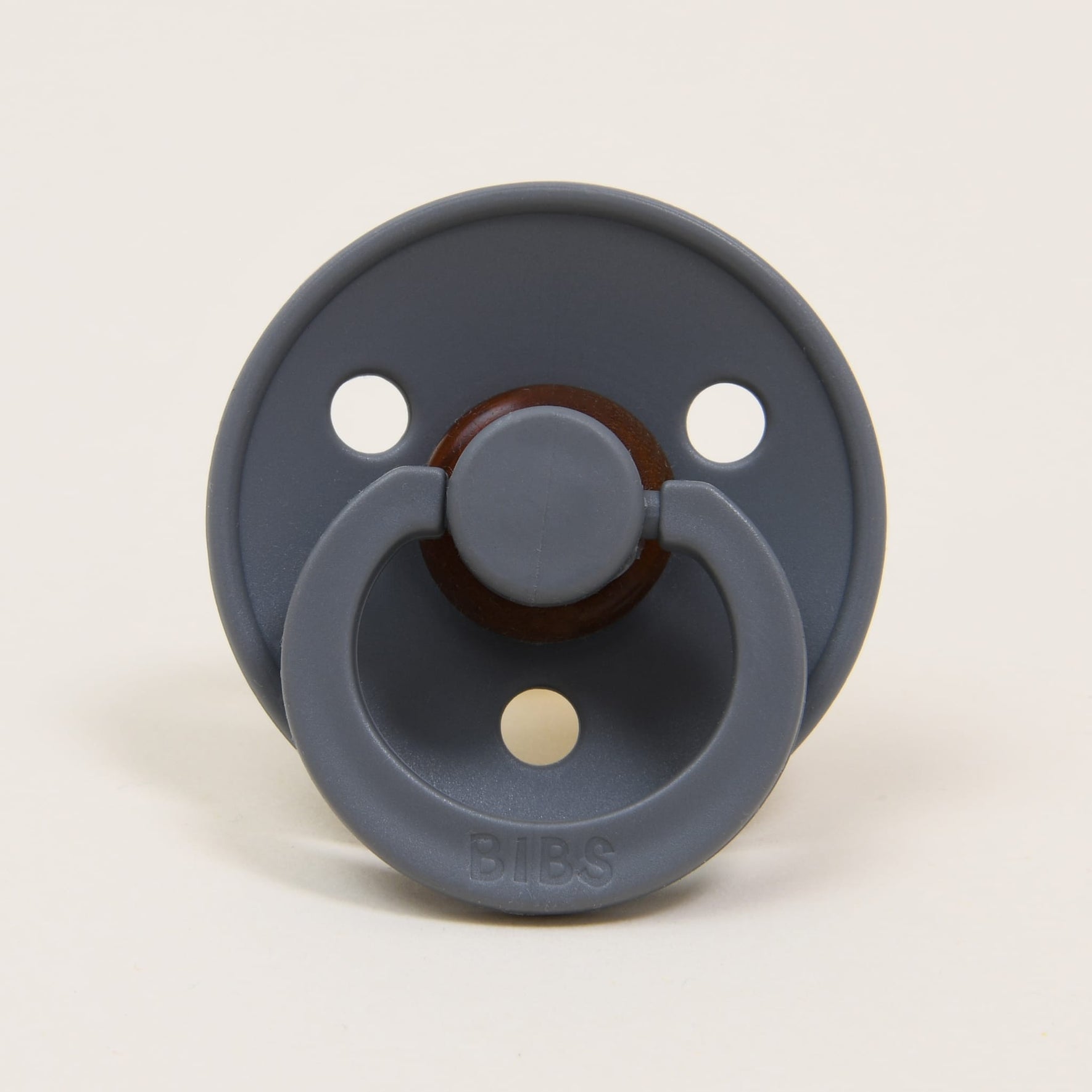 The Bibs Pacifier 2 Pack in Smoke features a natural rubber design made in Denmark, showcasing a dark gray round shield with three small holes and a central circular knob. The brand name "BIBS" is elegantly embossed on its lower part, ensuring a quality design that helps your little one self-soothe.