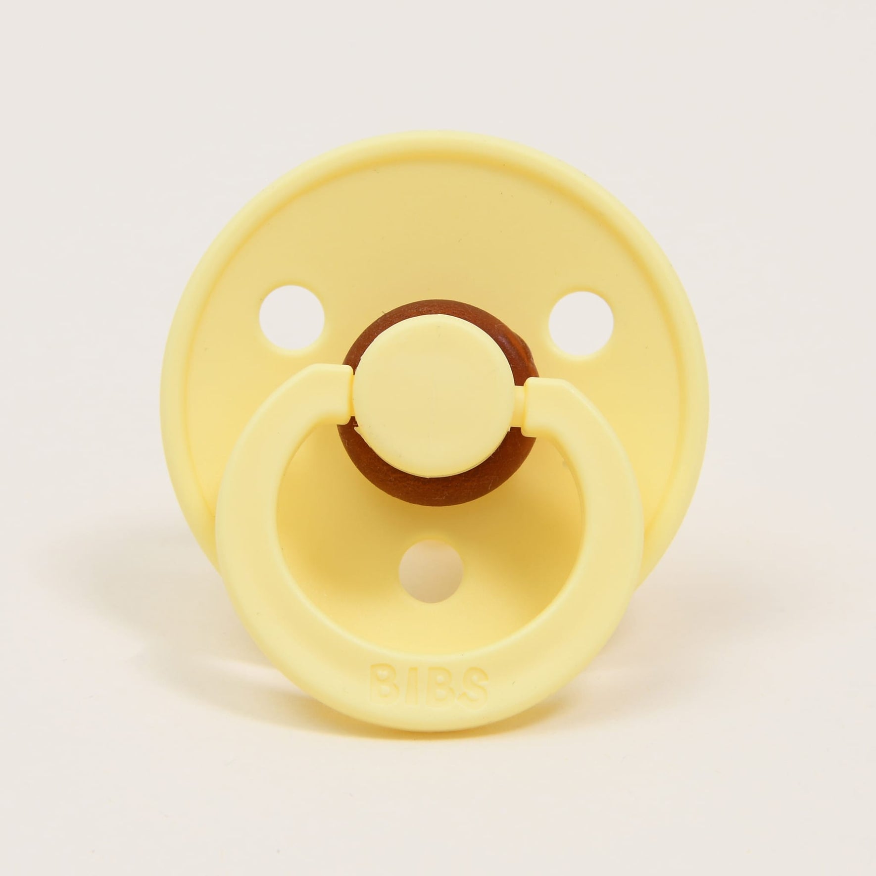 The "Bibs Pacifier 2 Pack | Sunshine" set includes a yellow pacifier made from natural rubber, featuring a classic design with a round shield and two holes on a plain white background. The central part and the ring are slightly darker in color, with the "BIBS" brand name embossed on the ring handle, ensuring comfort for babies as they self-soothe.