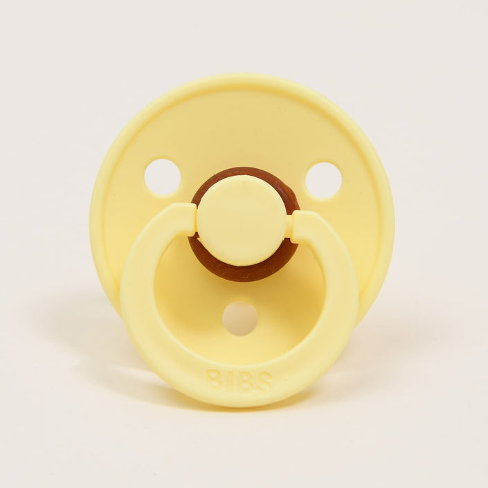 The "Bibs Pacifier 2 Pack | Sunshine" set includes a yellow pacifier made from natural rubber, featuring a classic design with a round shield and two holes on a plain white background. The central part and the ring are slightly darker in color, with the "BIBS" brand name embossed on the ring handle, ensuring comfort for babies as they self-soothe.