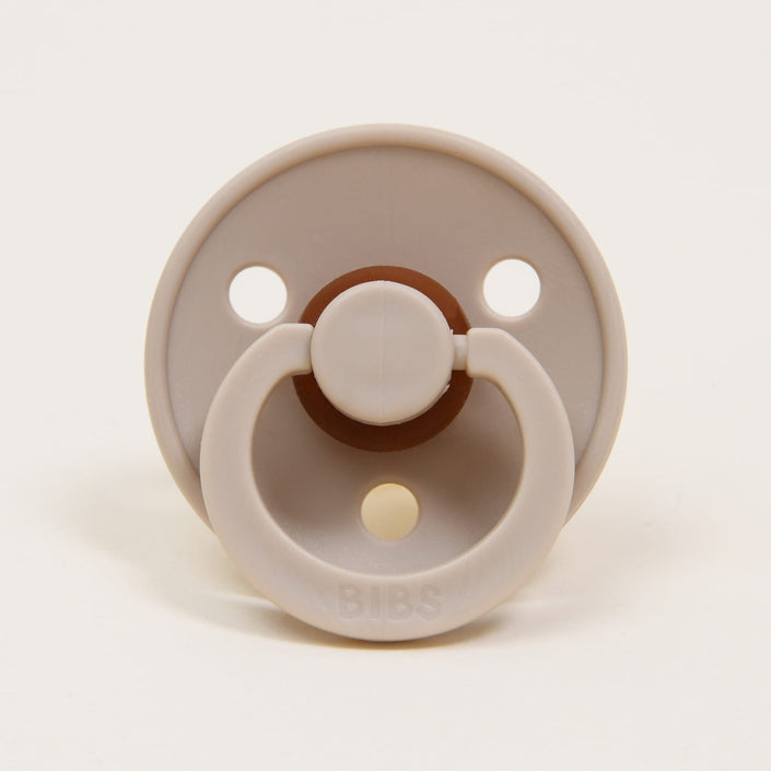 This beige Bibs Pacifier 2 Pack, manufactured in Denmark and named "Vanilla," showcases a round shield with three ventilation holes. Its classic look is accentuated by a ring handle and the "BIBS" logo embossed at the bottom. The nipple, crafted from natural rubber, boasts a contrasting darker hue that complements its sophisticated design.