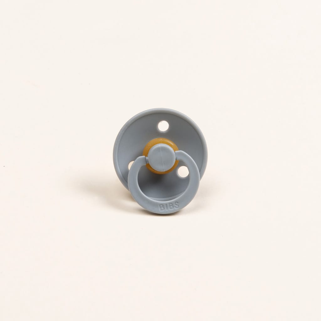 A single cloud-colored Bibs Pacifier, featuring a natural beige rubber nipple, is displayed against a simple off-white backdrop. This BPA-free pacifier includes two ventilation holes on its shield and has the brand name "BIBS" embossed on the handle, highlighting its quality as a product made in Denmark.