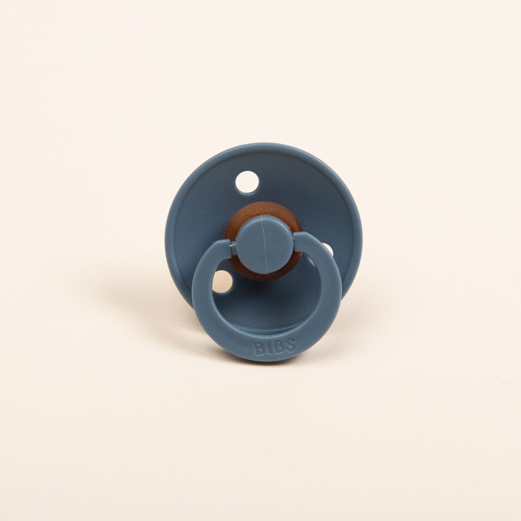 The image showcases a Bibs Pacifier, available in Petrol or Cloud colors, featuring a circular design with a brown natural rubber nipple. It is set against a simple, light-hued background. The BPA-free handle is embossed with the "BIBS" brand name, reflecting its Danish manufacturing that prioritizes safety and quality for your baby.