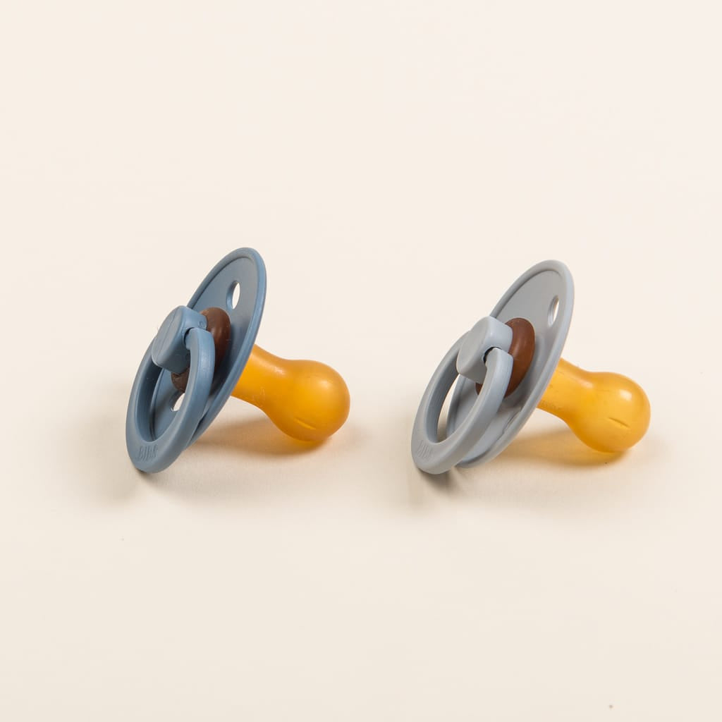 Two Bibs Pacifiers, made in Denmark, are displayed on a neutral background. Each pacifier features a yellow natural rubber teat and a light blue, BPA-free plastic shield in the "Petrol or Cloud" color option.