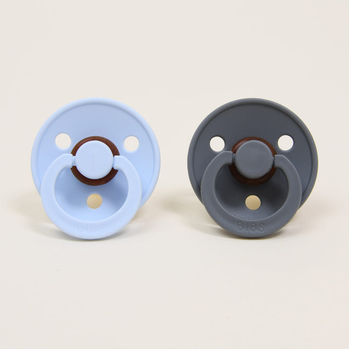 Displayed on a neutral backdrop, the Bibs Pacifier Set showcases two round pacifiers made from natural rubber. The pacifier to the left features a baby blue hue, while the one on the right is in smoke gray. Both sport circular shields and ring handles, providing a calming option for infants.