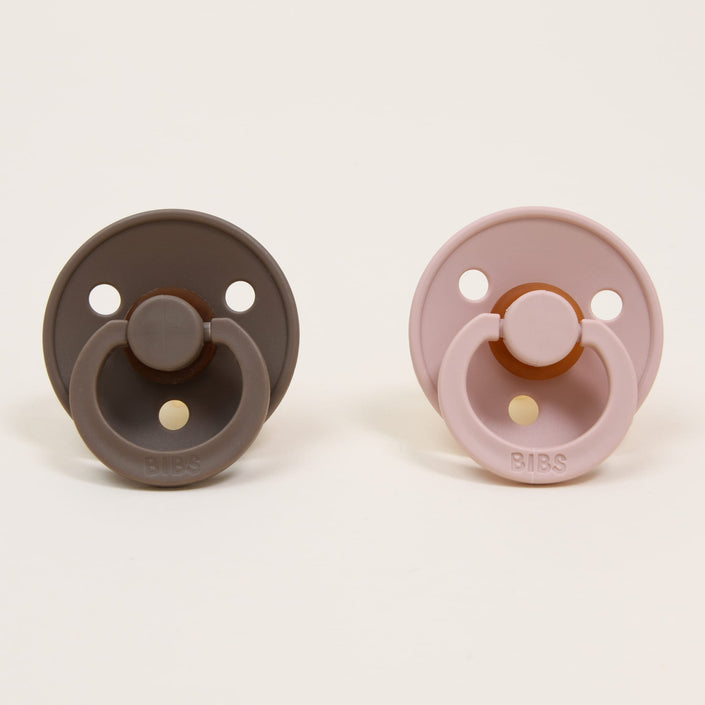 The Bibs Pacifier Set from Denmark features two pacifiers displayed side by side. The one on the left, crafted from natural rubber and called Dark Oak, is a dark brown shade, while the right one is Light Pink, known as Blush. Both have a simple round design with a looped handle, ideal for helping babies self-soothe against a plain background.