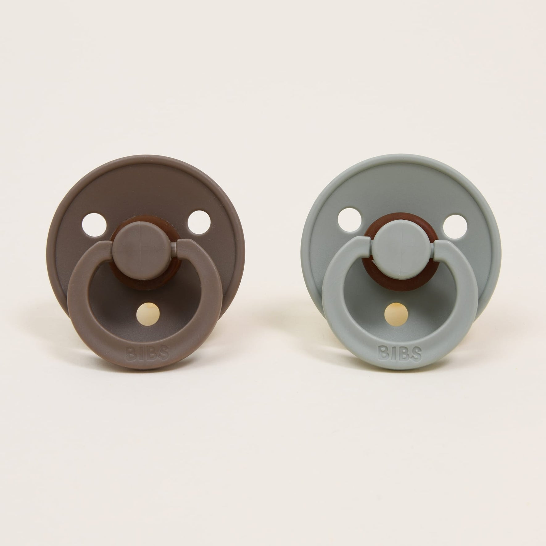 Two Bibs pacifiers from the Dark Oak & Sage set are displayed against a plain background. The classic design features the pacifier on the left in a dark oak shade and the one on the right in sage. Both have round shields with two ventilation holes and a ring handle, ideal for self-soothing.