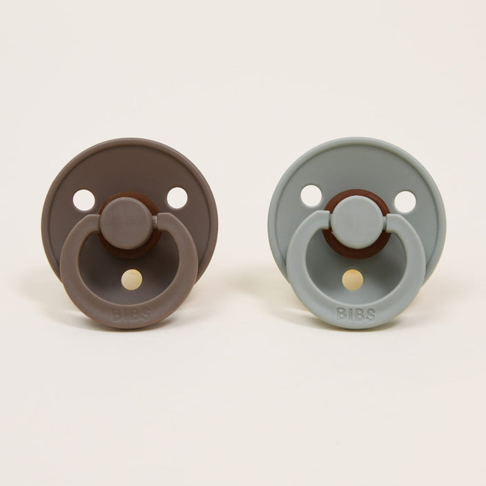 Two Bibs pacifiers from the Dark Oak & Sage set are displayed against a plain background. The classic design features the pacifier on the left in a dark oak shade and the one on the right in sage. Both have round shields with two ventilation holes and a ring handle, ideal for self-soothing.