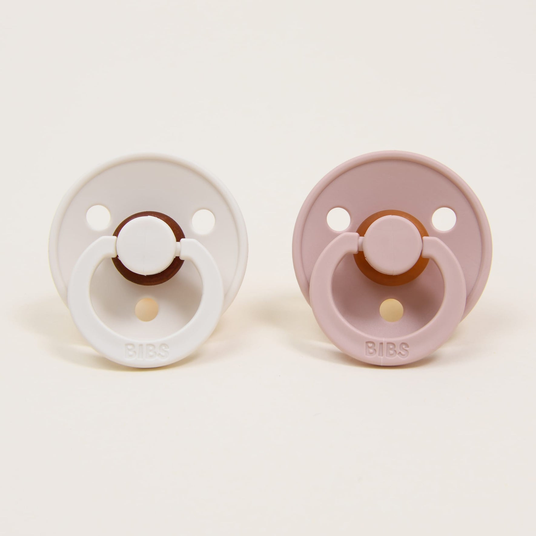 The Bibs Pacifier Set, featuring two round baby pacifiers with a classic design, is displayed against a plain background. The ivory natural rubber pacifier on the left contrasts with the blush self-soothe option on the right. Both pacifiers include a circular guard and handle.