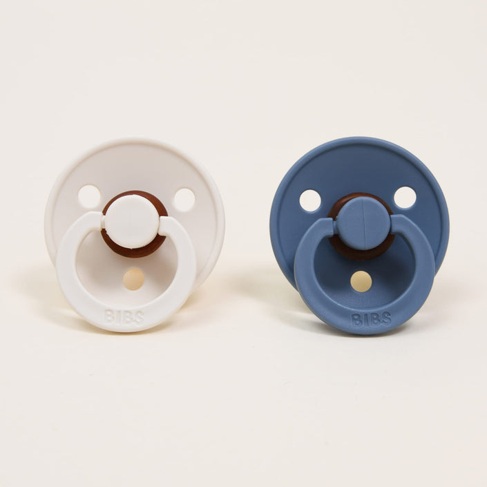 The Bibs Pacifier Set in Ivory and Petrol features two pacifiers displayed against a plain background. Made in Denmark, the one on the left is ivory, while the one on the right is petrol blue. Both pacifiers are crafted from natural rubber and share a similar round shape, complete with two ventilation holes and a ring handle for self-soothing. The brand name "BIBS" is embossed on each ring.
