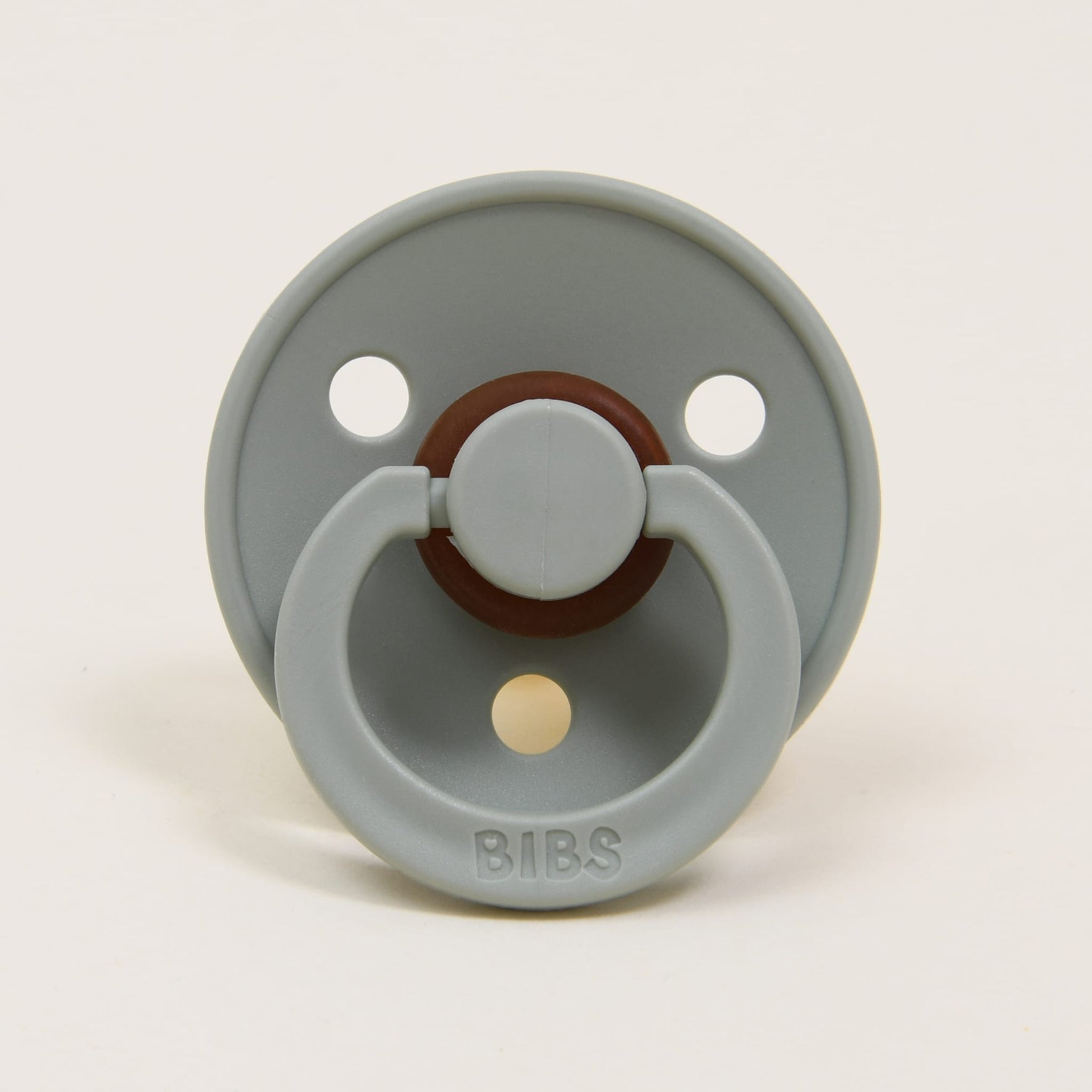 An ivory BIBS pacifier from the Ivory & Sage Set, made in Denmark, displays a round shield with three ventilation holes. Constructed from natural rubber, it includes a dark brown central ring and an arched handle, ideal for assisting your baby in self-soothing. The pacifier is shown head-on against a simple background.