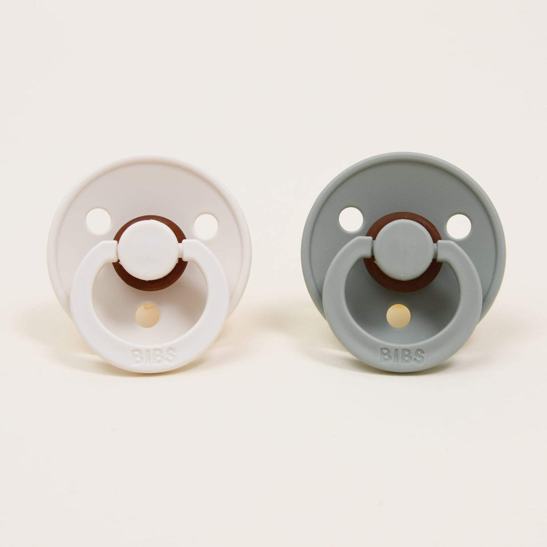 Against a plain backdrop, two pacifiers from the Bibs Pacifier Set in Ivory & Sage are displayed, showcasing their sleek design. These options feature round shields with ventilation holes and rings adorned with the "BIBS" brand logo. Made in Denmark from natural rubber, these pacifiers are ideal for helping babies self-soothe.