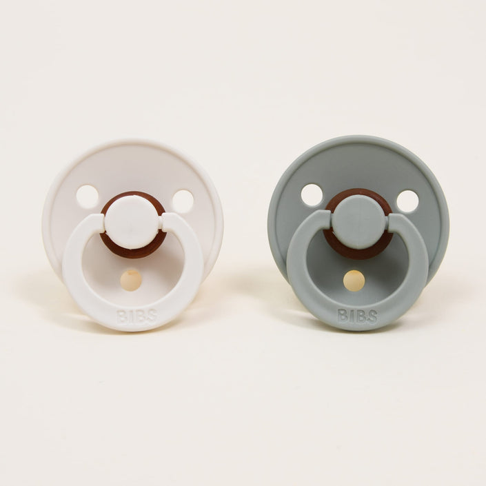 Against a plain backdrop, two pacifiers from the Bibs Pacifier Set in Ivory & Sage are displayed, showcasing their sleek design. These options feature round shields with ventilation holes and rings adorned with the "BIBS" brand logo. Made in Denmark from natural rubber, these pacifiers are ideal for helping babies self-soothe.
